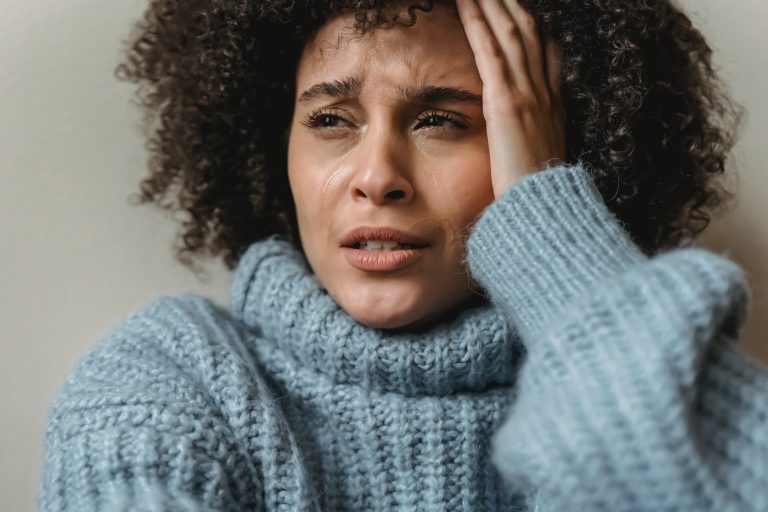 Woman is sitting against a wall, crying with a hand to her face