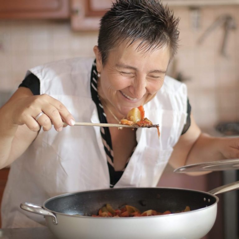 a woman smiles as she bends over the pan to taste the food on the spoon, slightly self conscious of the photo but fully confident in the kitchen