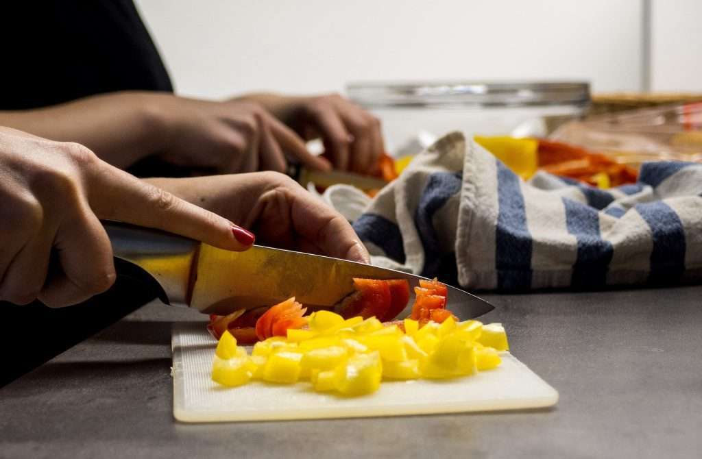 Two women's hands are visible, confidently chopping bell peppers together on a kitchen counter.