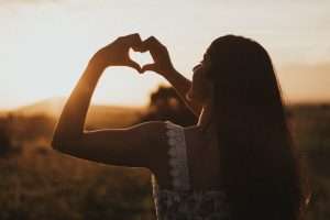 a woman holds her hands in a heart shape toward the sunset