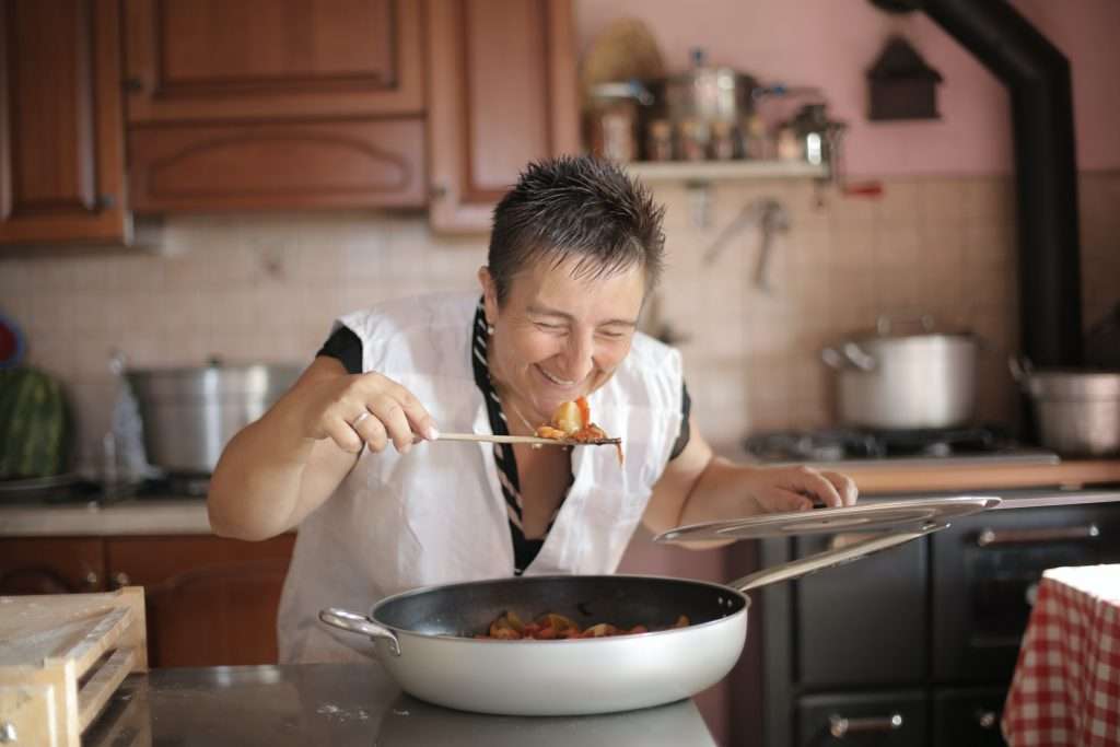 a woman smiles as she bends over the pan to taste the food on the spoon, slightly self conscious of the photo but fully confident in the kitchen