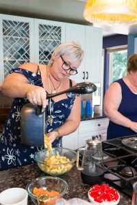 A woman pours colorful pasta from a pot into a bowl, while another woman cooks something else nearby