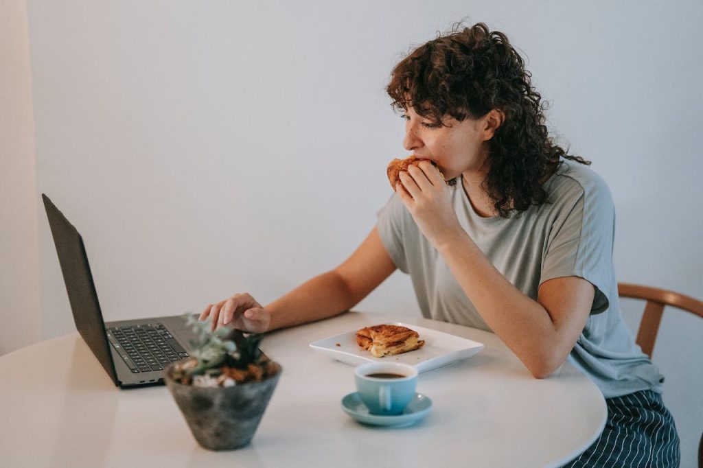 Woman sits at table, eating a sandwich and working on laptop